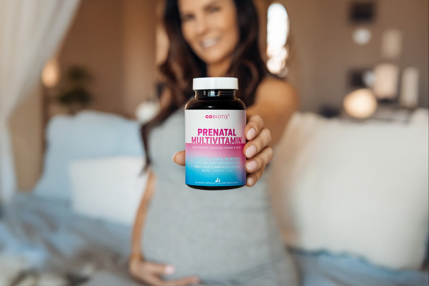 Pregnant woman holding a bottle of GoBiotix Prenatal Multivitamin in a bedroom setting.